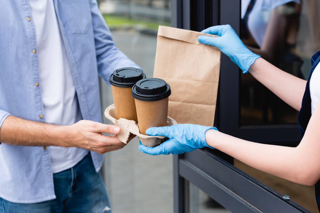Widok człowieka odbierającego paczkę i papierowe kubki od kelnerki w rękawiczkach lateksowych przy wejściu do kawiarni  - Zdjęcie, obraz
