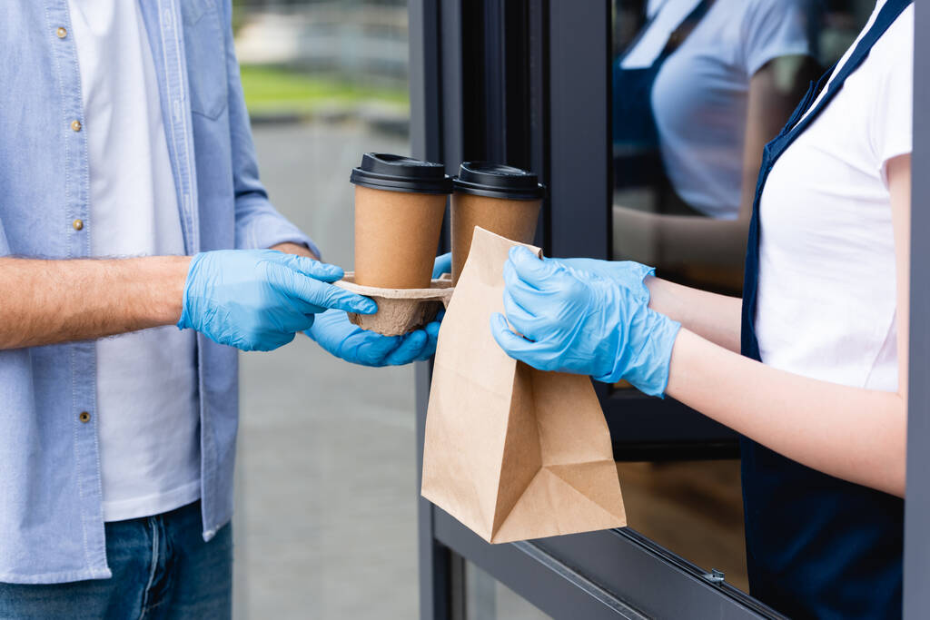 Ausgeschnittene Ansicht eines Mannes, der Pappbecher in der Hand hält und Pakete von einer Kellnerin in Latexhandschuhen in der Nähe eines Cafés entgegennimmt  - Foto, Bild
