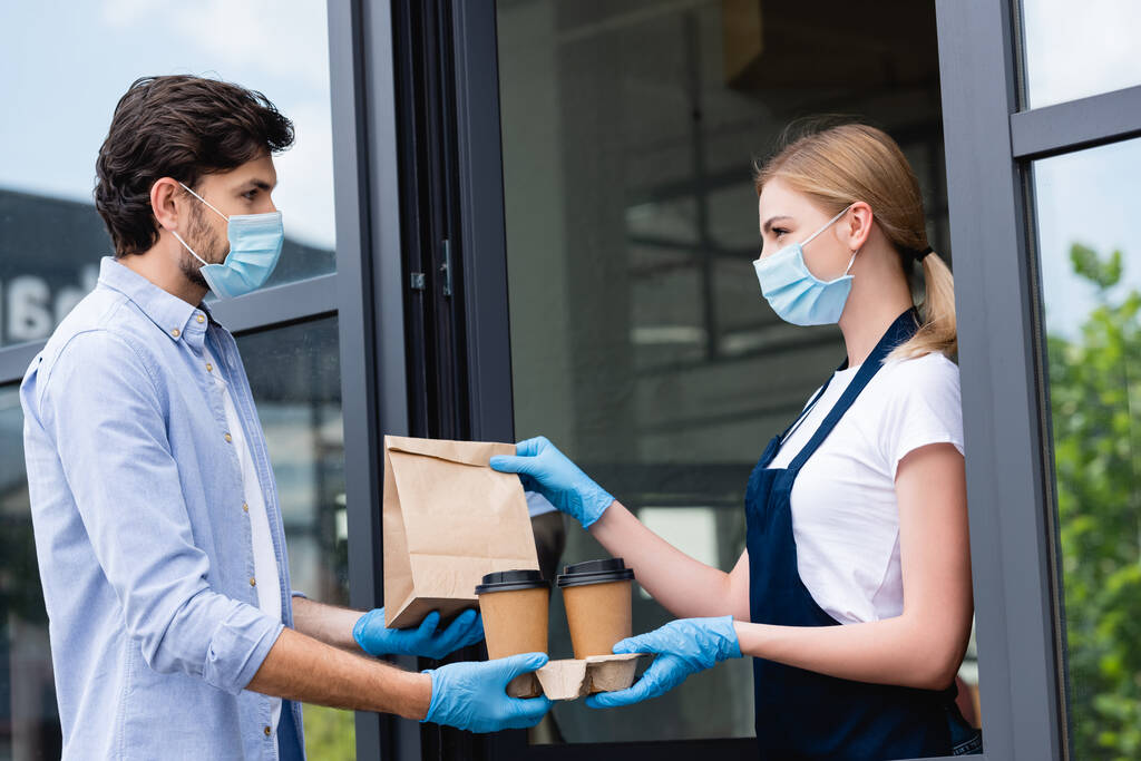 Widok z boku człowieka w lateksowych rękawiczkach i maskach medycznych otrzymujących zamówienie od kelnerki w pobliżu kawiarni - Zdjęcie, obraz