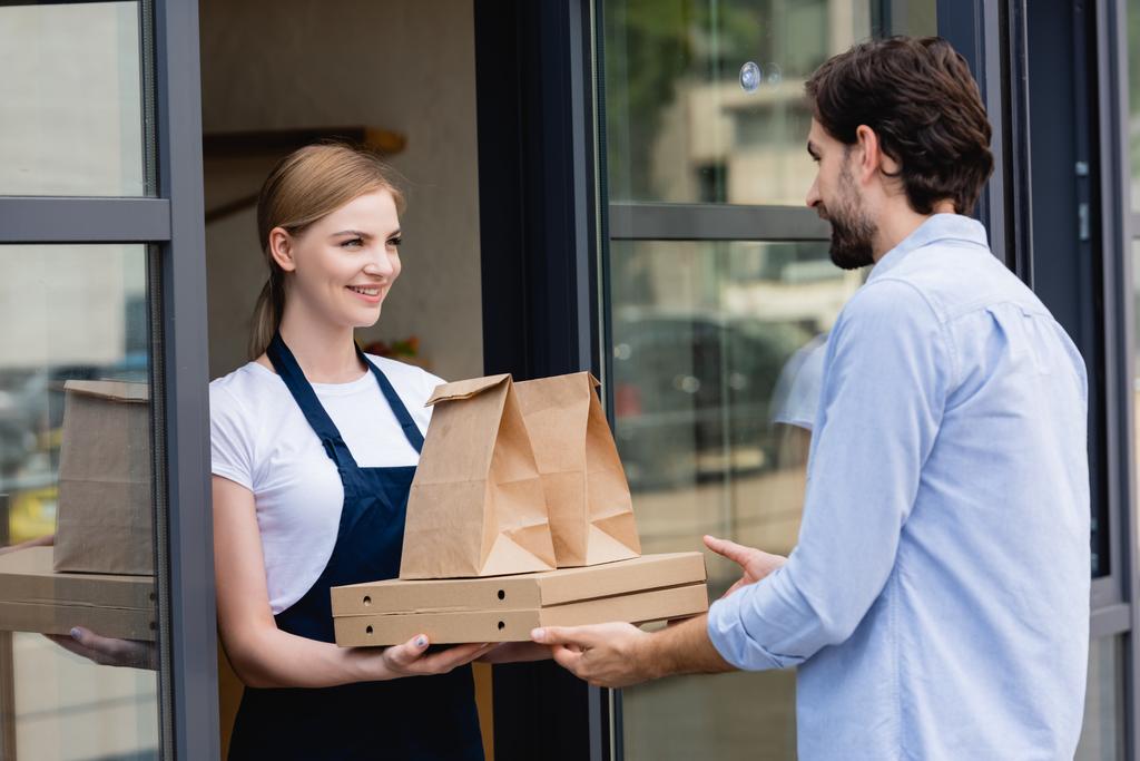 Atractiva camarera dando cajas de pizza y bolsas de papel al hombre en la entrada de la cafetería
  - Foto, imagen