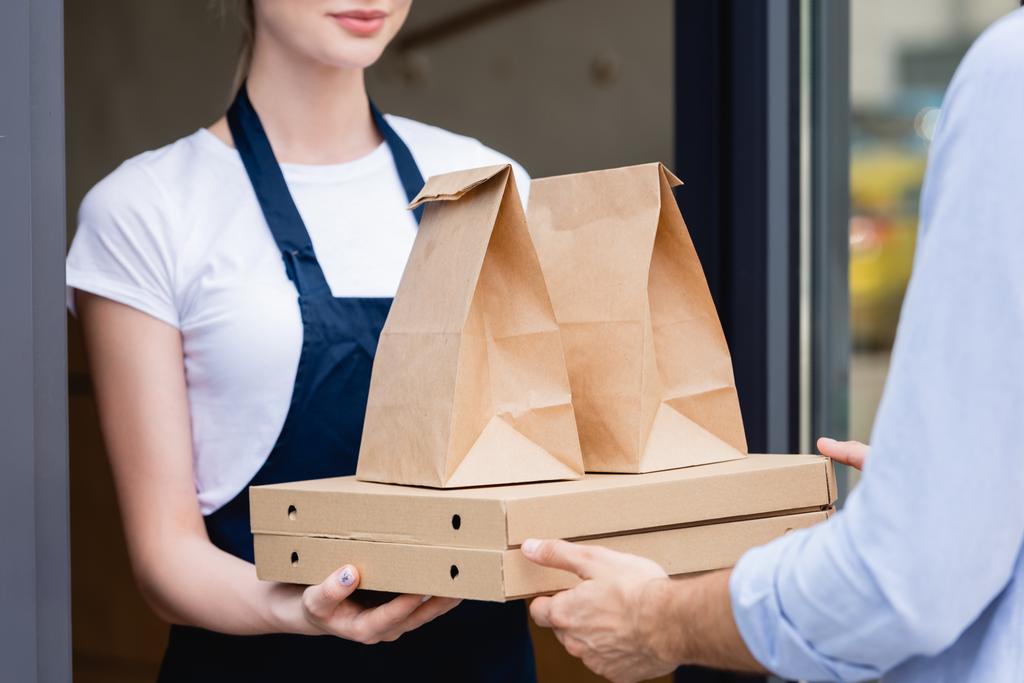 Обрезанный вид на человека, получающего коробки пиццы и пакеты от официантки возле кафе  - Фото, изображение