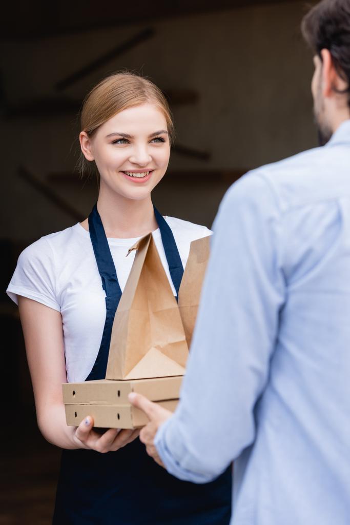 Enfoque selectivo de la hermosa camarera mirando al hombre y dando cajas de pizza y bolsas de papel en la entrada del edificio
  - Foto, imagen
