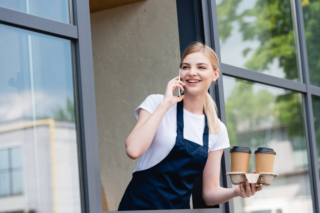 Low angle view of smiling waitress talking on smartphone and holding paper cups near window of cafe - Photo, Image