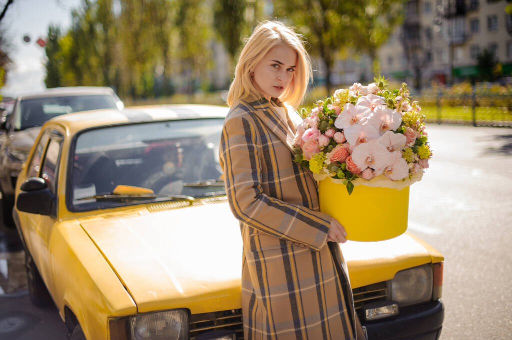mladá blondýnka drží kulatou žlutou krabici s květinovým uspořádáním růží a orchidejí a dívá se do kamery - Fotografie, Obrázek