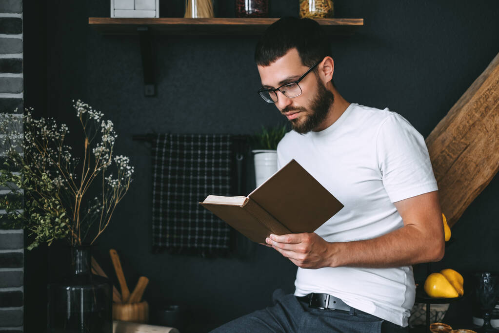 Een jonge blanke man met een baard die een boek leest in de keuken. Thuis vrije tijd. Hoge kwaliteit foto - Foto, afbeelding