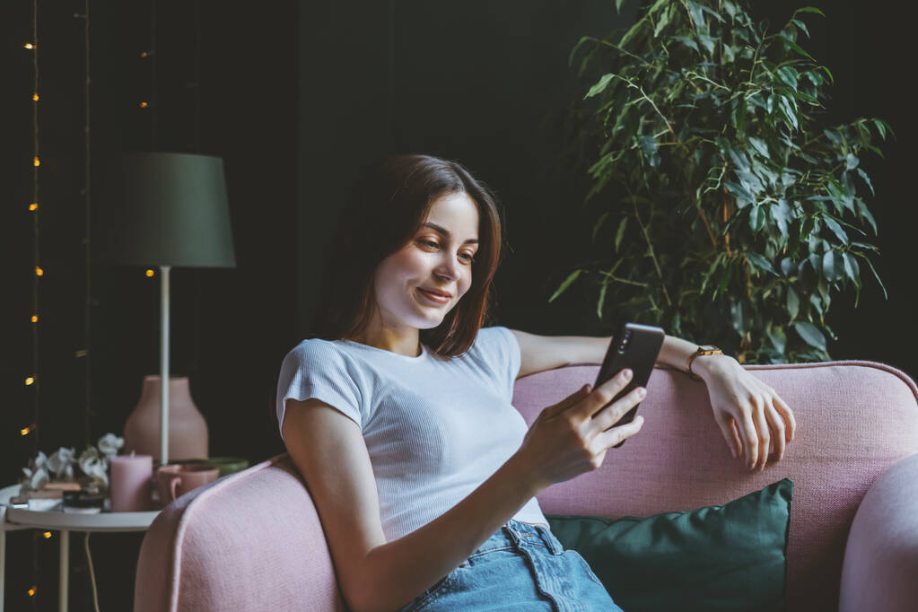 Młoda, uśmiechnięta biała kobieta używa smartfona siedząc na różowej kanapie w domu. Wysokiej jakości zdjęcie - Zdjęcie, obraz