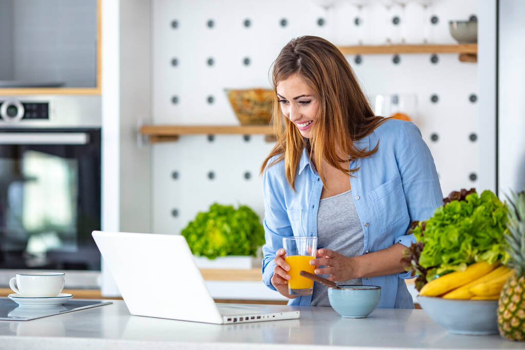 Kaunis nuori nainen työskentelee kannettavan tietokoneen kanssa syödessään keittiössä. Viehättävä nuori nainen surffaamassa internetissä kotona. Aina netissä. Nainen surffaa netissä nauttien aamiaista keittiössään kotona - Valokuva, kuva