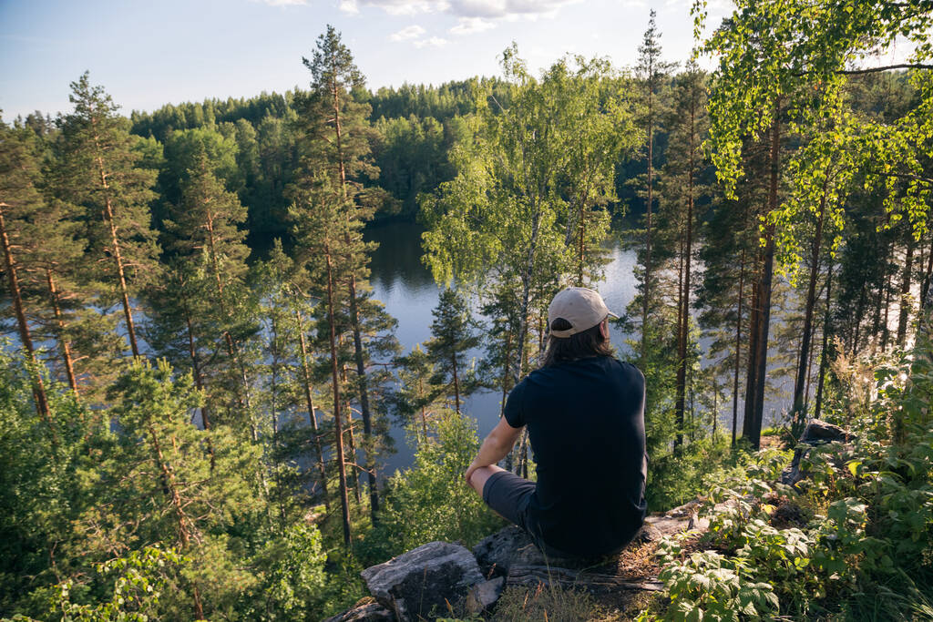 Samiec podróżujący w czapce siedzi na krawędzi klifu i spogląda w oddali do lasu. Piękny krajobraz, podróże i koncepcja turystyki. - Zdjęcie, obraz