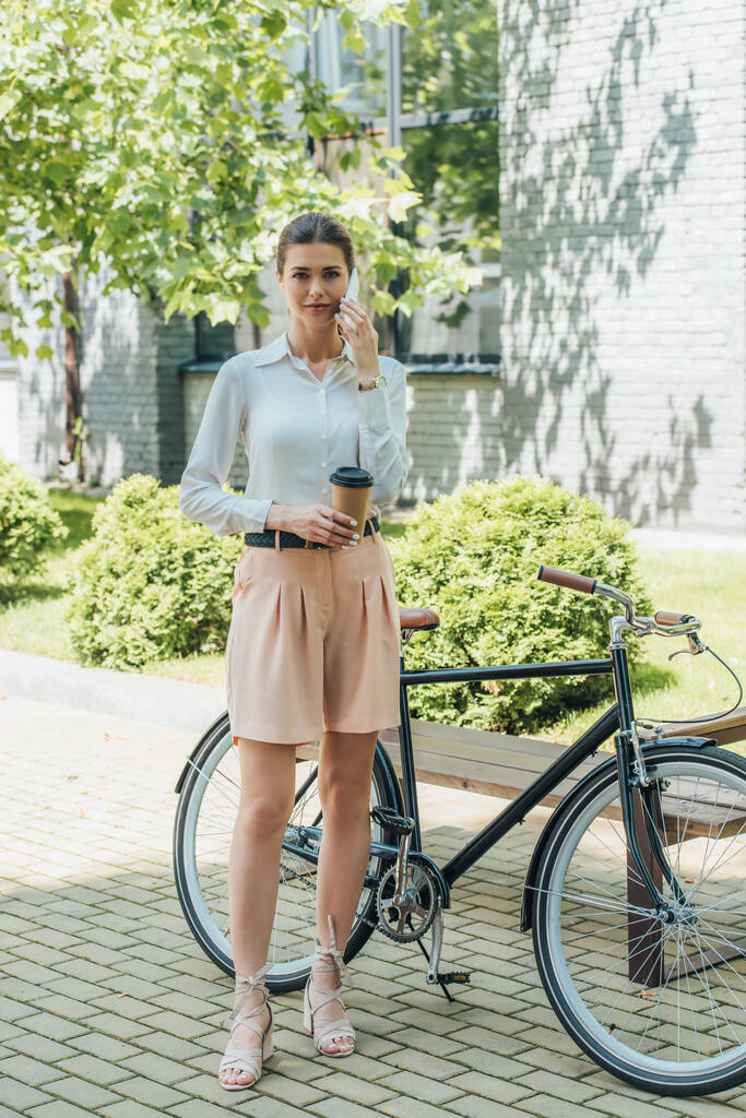 attraktive Geschäftsfrau spricht mit Smartphone und hält Pappbecher in der Nähe von Fahrrad  - Foto, Bild