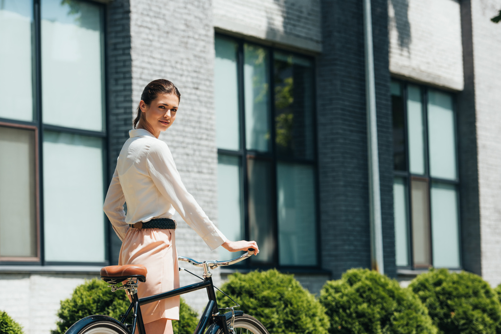 attractive businesswoman walking with bicycle near modern building  - Photo, Image
