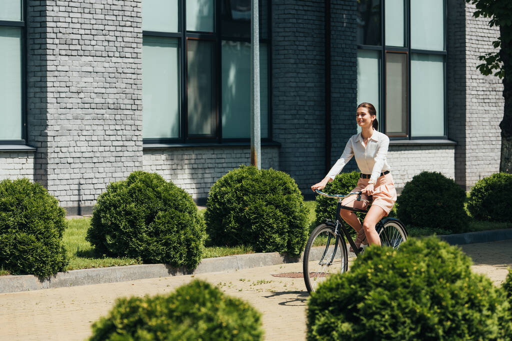 selectieve focus van gelukkige zakenvrouw paardrijden fiets in de buurt van modern gebouw  - Foto, afbeelding