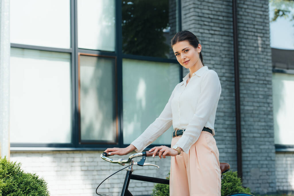 mooie jonge zakenvrouw staan met fiets in de buurt van modern gebouw  - Foto, afbeelding