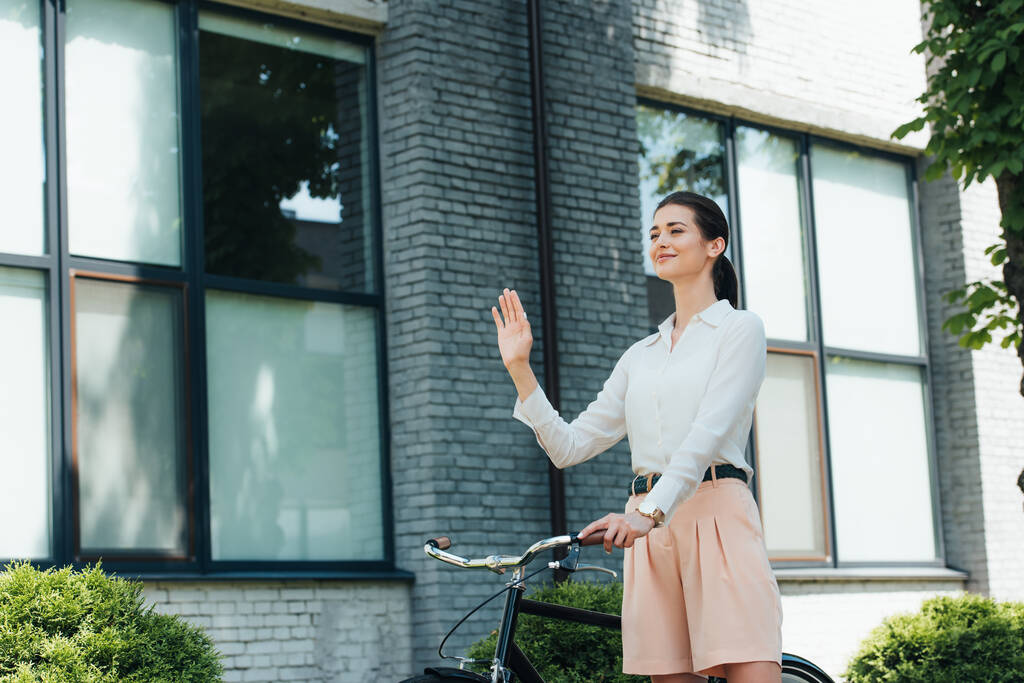 radosna młoda bizneswoman stojąca na rowerze i machająca ręką  - Zdjęcie, obraz