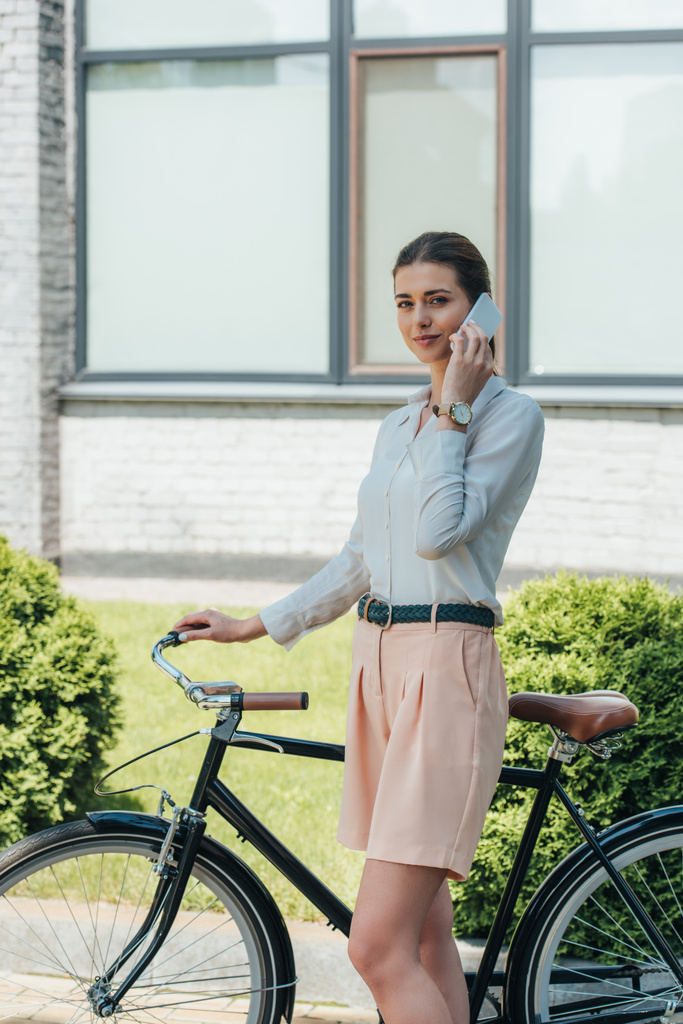 aantrekkelijke zakenvrouw praten op smartphone terwijl staan in de buurt van fiets en gebouw  - Foto, afbeelding