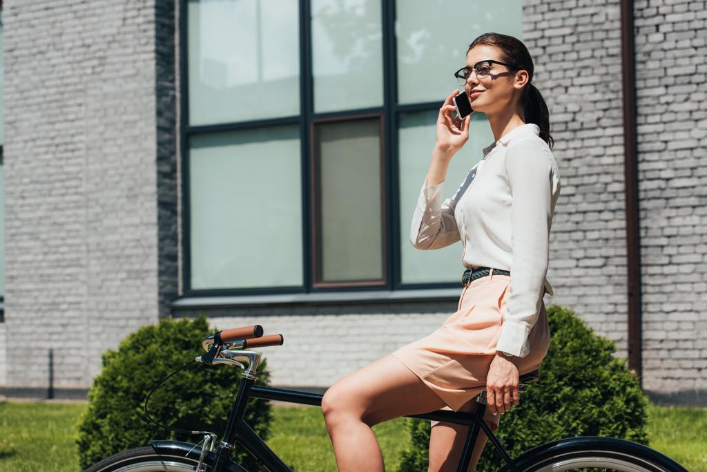attraktive Geschäftsfrau mit Brille, die auf dem Smartphone spricht und auf dem Fahrrad sitzt  - Foto, Bild