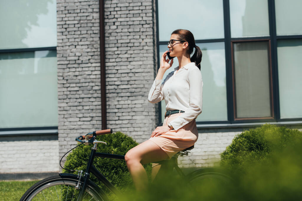 selectieve focus van zakenvrouw in bril praten op smartphone en zitten op de fiets  - Foto, afbeelding