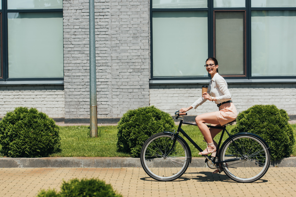 selective focus of cheerful businesswoman in glasses riding bicycle and holding paper cup - Photo, Image