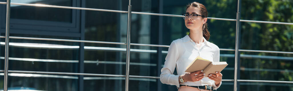 culture panoramique de femme d'affaires dans des lunettes tenant livre près du bâtiment moderne  - Photo, image