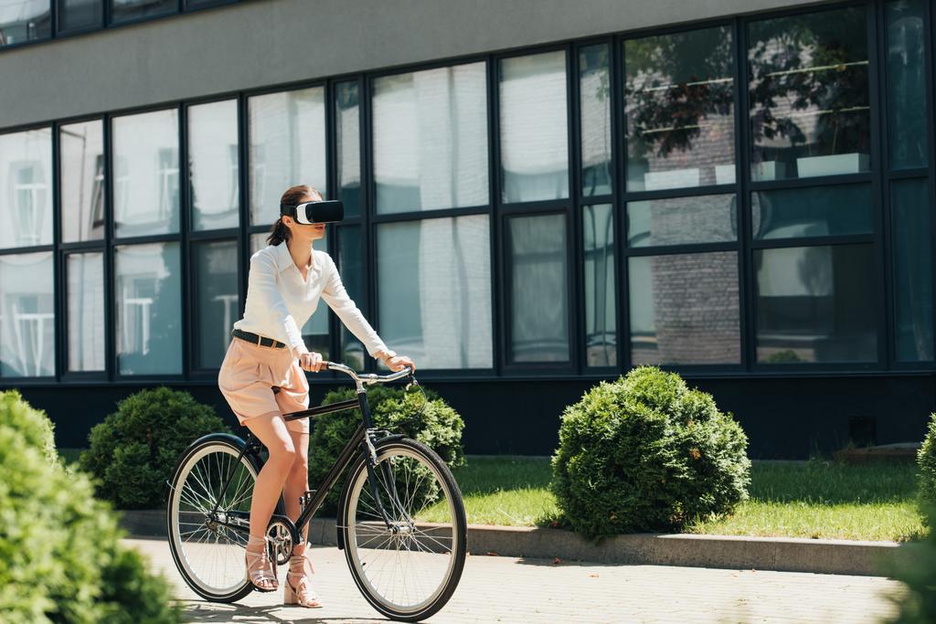 selectieve focus van jonge zakenvrouw in virtual reality koptelefoon paardrijden fiets in de buurt van gebouw  - Foto, afbeelding