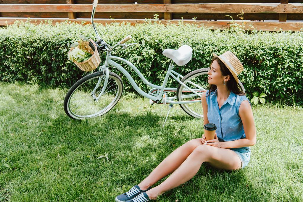 γυναίκα σε ψάθινο καπέλο κρατώντας χάρτινο κύπελλο και κάθεται στο γρασίδι κοντά στο ποδήλατο  - Φωτογραφία, εικόνα