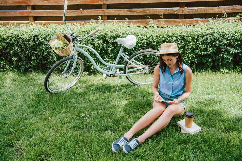 κορίτσι σε ψάθινο καπέλο χρησιμοποιώντας ψηφιακό tablet και κάθεται στο γρασίδι κοντά στο ποδήλατο, χάρτινο κύπελλο και βιβλία - Φωτογραφία, εικόνα