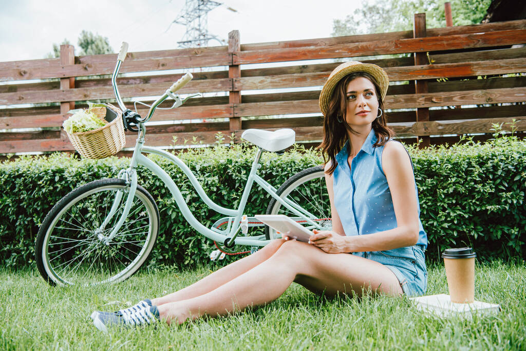 mulher de chapéu de palha segurando tablet digital e sentado na grama perto de bicicleta, copo de papel e livro
 - Foto, Imagem