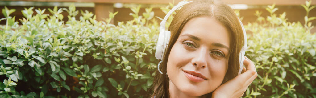 plan panoramique de jeune femme joyeuse écoutant de la musique dans des écouteurs sans fil près de la brousse  - Photo, image