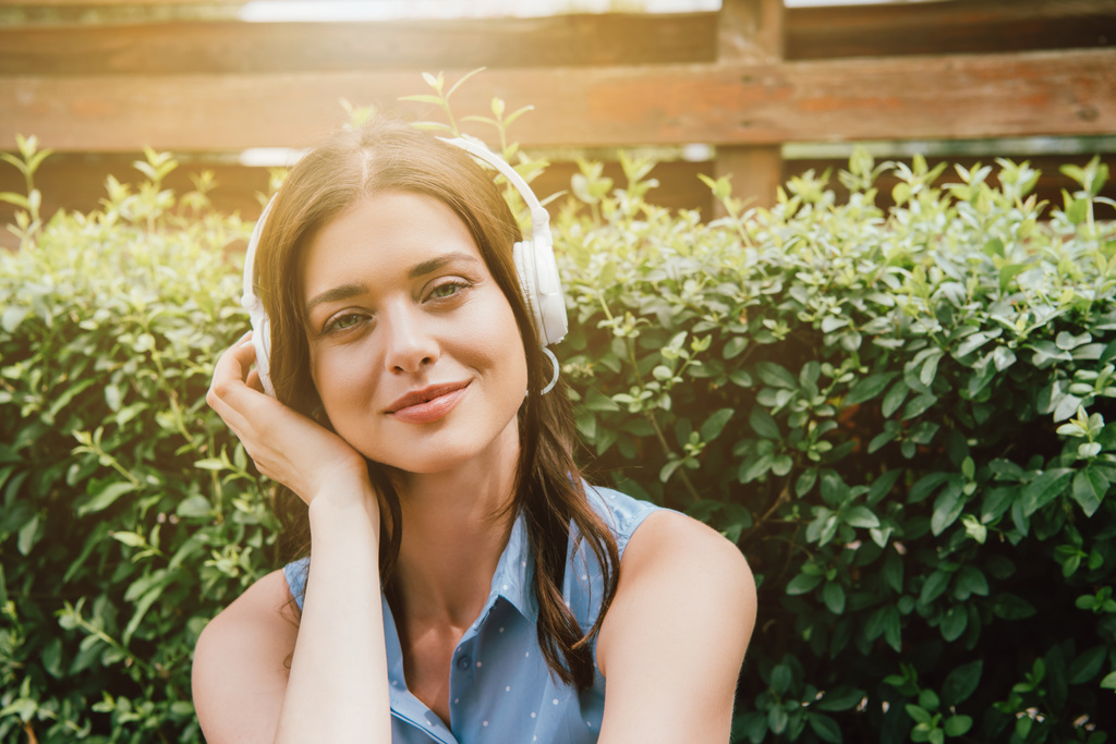 glückliche Frau hört Musik in drahtlosen Kopfhörern in der Nähe von Busch  - Foto, Bild