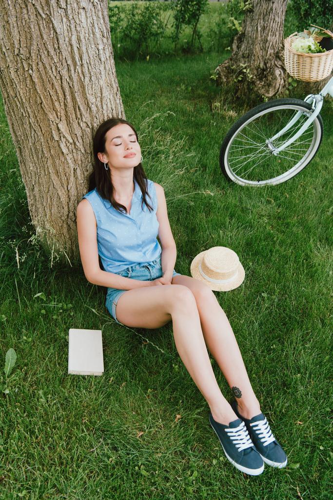 vue grand angle de femme heureuse assise sur l'herbe près du tronc d'arbre, vélo, livre et chapeau de paille  - Photo, image