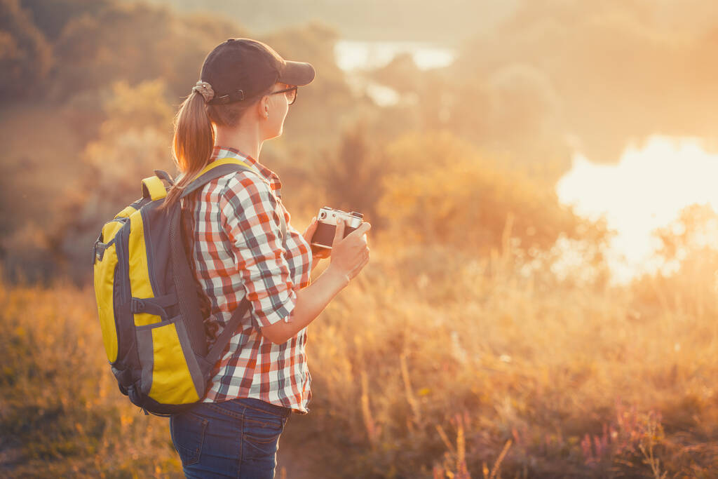 tourist girl with a backpack and a camera walks through a picturesque meadow and photographs the picturesque landscape. Tourism, outdoor activities - Photo, Image