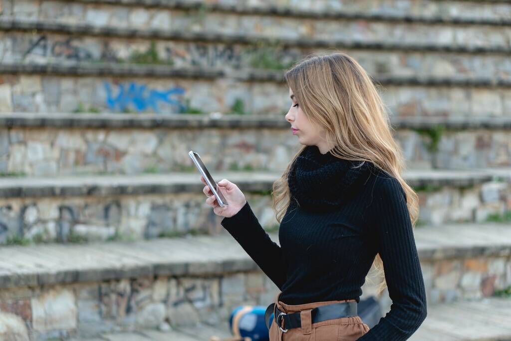 Een jonge blonde vrouw in een zwarte coltrui en beige broek staart naar haar telefoon - Foto, afbeelding