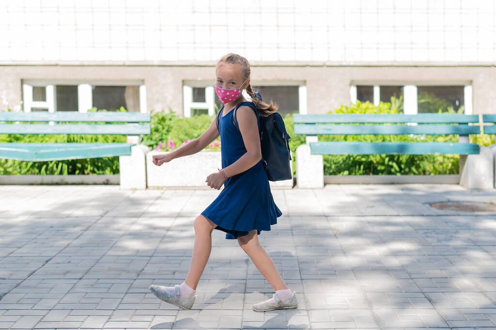 Joyful little girl in a blue dress and a protective mask runs to school. A happy schoolgirl with a pigtail and a backpack on her back runs into a bounce to classes. Mask mode. - Photo, Image