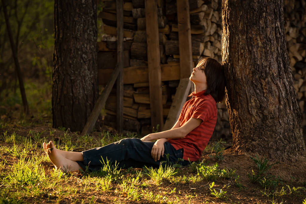 Kırmızı tişörtlü bir çocuk gün ışığında çimlerin üzerinde oturur. - Fotoğraf, Görsel