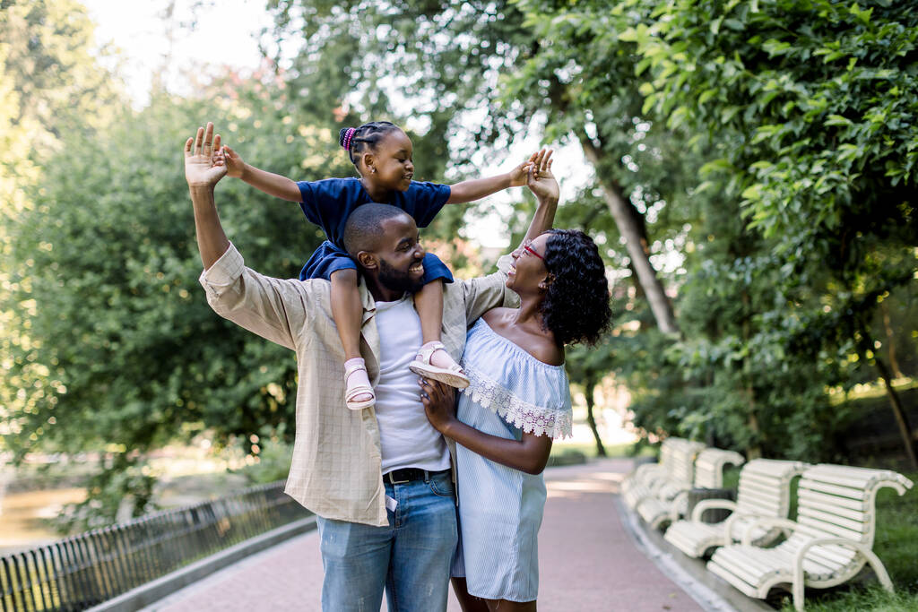 Młoda szczęśliwa para afrykańskich rodziców, spędzająca razem czas i ciesząca się ciepłym letnim wieczorem w parku, spacerująca ze swoją małą uroczą córką, siedząca na ramionach ojca - Zdjęcie, obraz