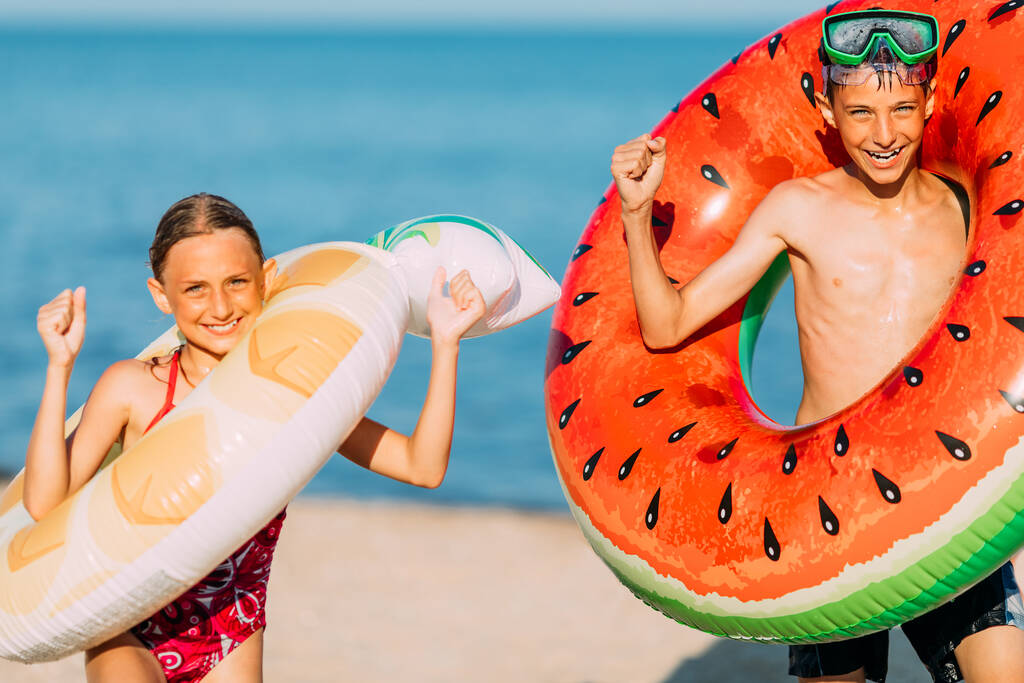 Enfants heureux un petit garçon et une fille s'amusent sur la plage avec des cercles de natation gonflables, montrent un geste gagnant. Voyages, vacances d'été - Photo, image