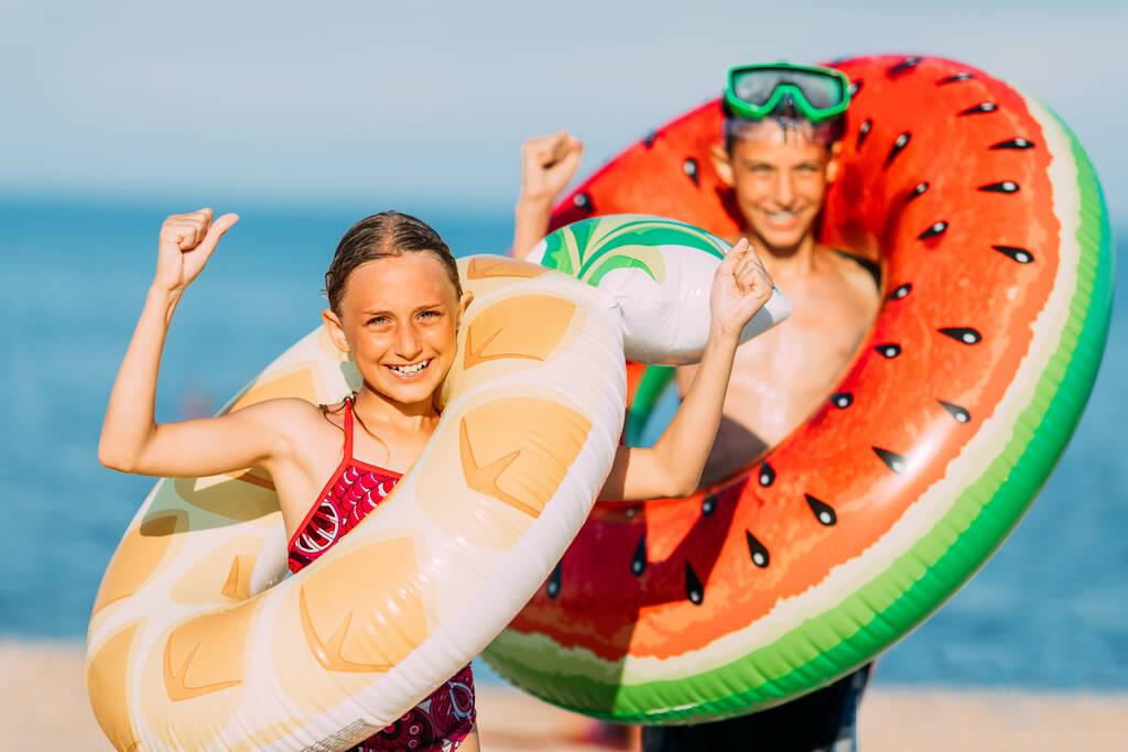 Szczęśliwe dzieci mały chłopiec i dziewczynka bawić się na plaży z nadmuchiwane koła pływackie, pokazać zwycięski gest. Podróże, letnie wakacje - Zdjęcie, obraz