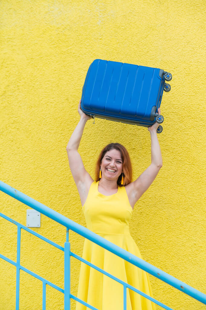 Donna felice in un abito giallo tiene una valigia blu sopra la testa sullo sfondo di una parete gialla. Ragazza caucasica sorride con i denti e aspetta felicemente un viaggio. - Foto, immagini
