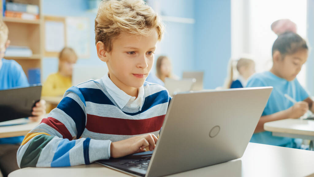 Escola Primária de Ciência da Computação Classe: Rapaz Inteligente Usa Computador Laptop, Seus colegas de classe trabalham com laptops também. Crianças obtendo educação moderna em STEM, brincando e aprendendo
 - Foto, Imagem