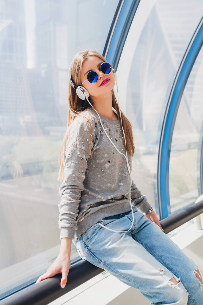 молода хіпстерка в повсякденному вбранні весело слухає музику в навушниках, студент у відпустці, носить джинси, светр і сонцезахисні окуляри, міський стиль
 - Фото, зображення