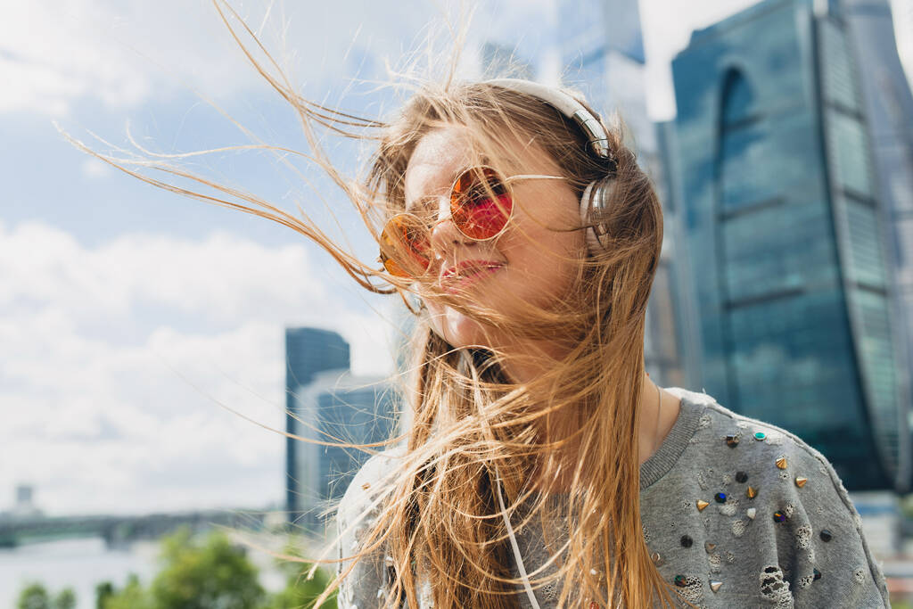 joven mujer hipster divertirse en la calle escuchando música en los auriculares, con gafas de sol de color rosa, estudiante de vacaciones, primavera verano estilo urbano, pelo largo y rubio
 - Foto, imagen