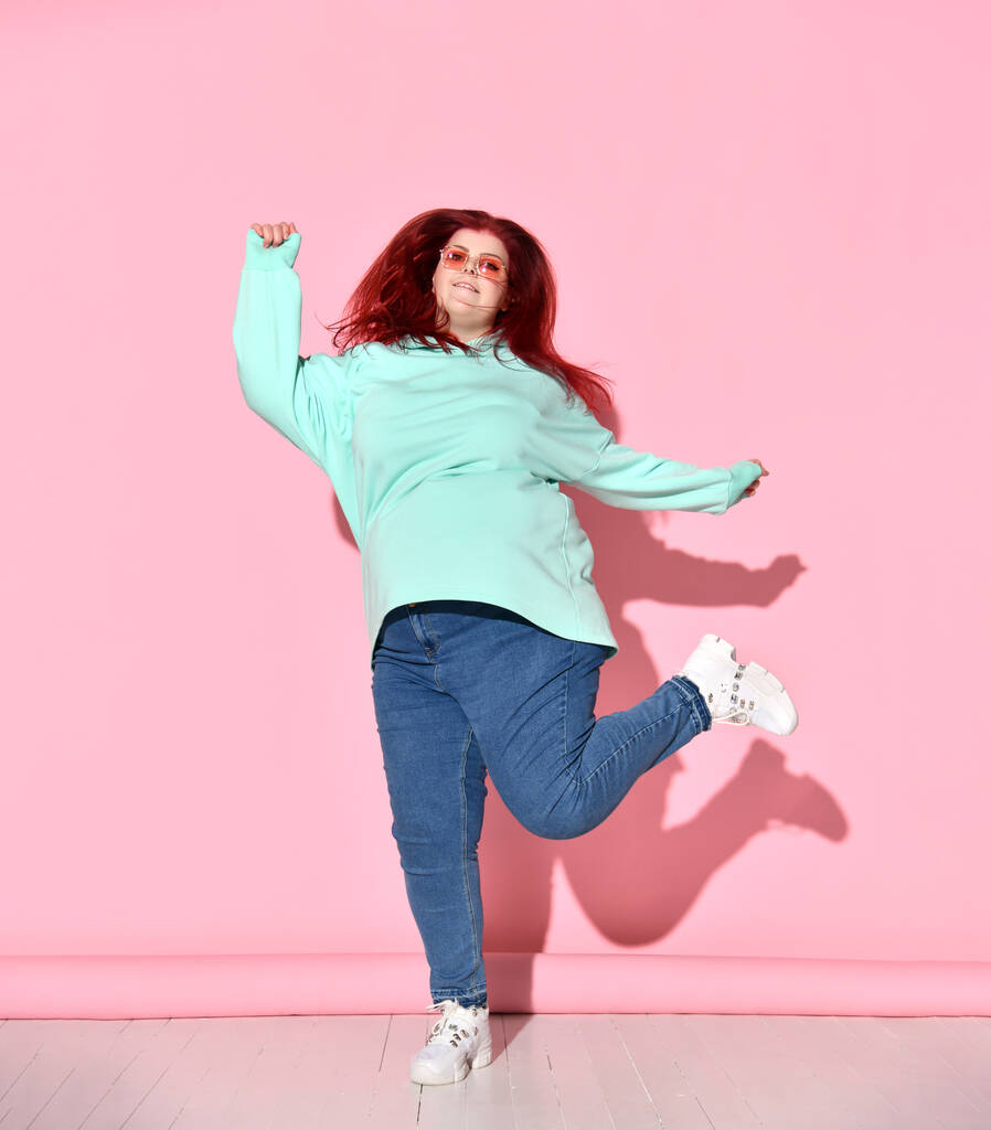 Mujer bastante regordeta con pelo de jengibre en jeans, sudadera con capucha acogedora y zapatillas de deporte que se divierten mucho saltando en el suelo, aislado en rosa
 - Foto, imagen