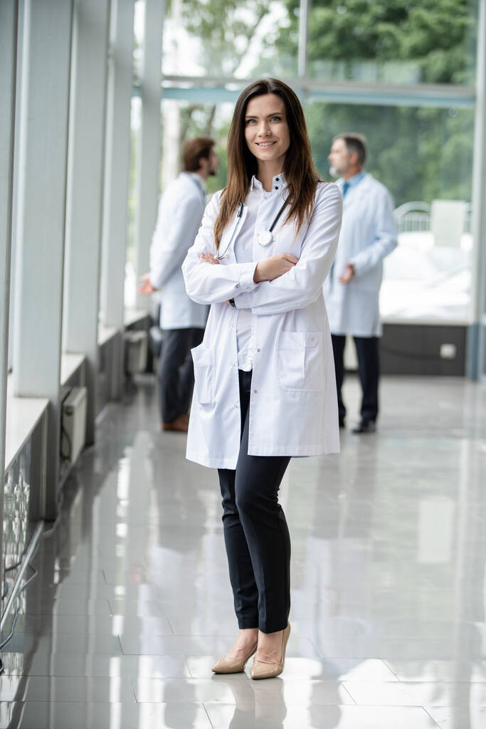 Retrato de una joven doctora con una bata blanca de pie en el hospital. - Foto, Imagen