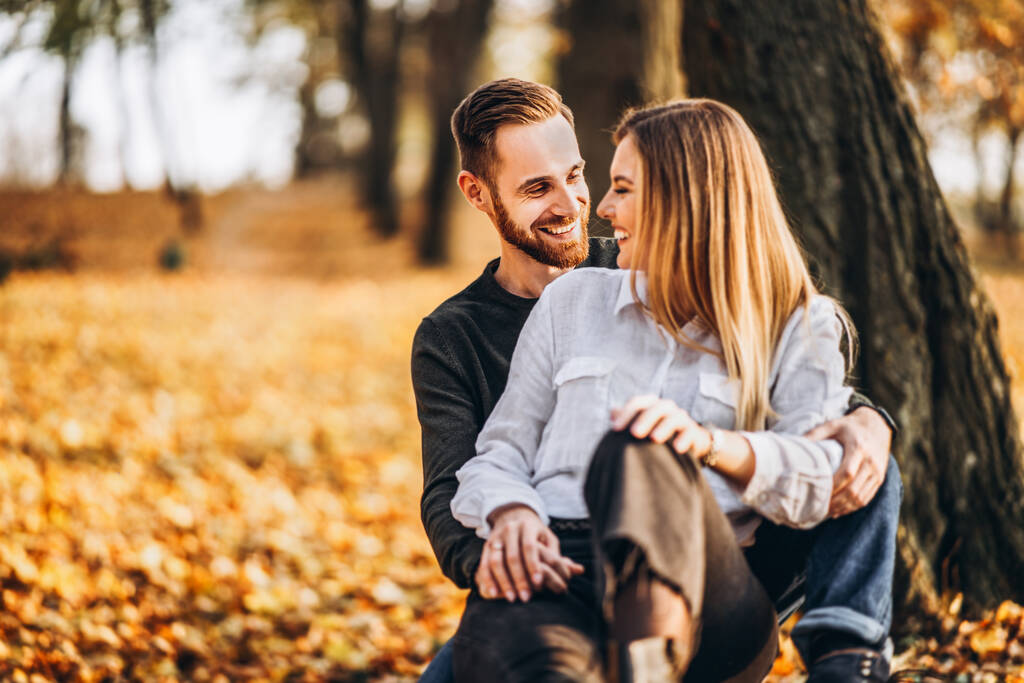 Молода закохана пара сидить на дерев'яній лавці в лісі. Чоловік і жінка обіймаються і посміхаються на тлі осінніх дерев
. - Фото, зображення