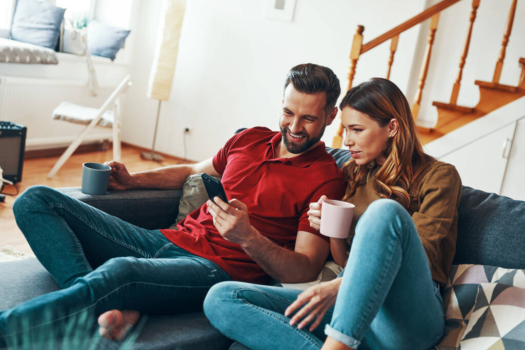 Happy young couple in casual clothing bonding together and smiling while resting on the sofa indoors - Photo, Image