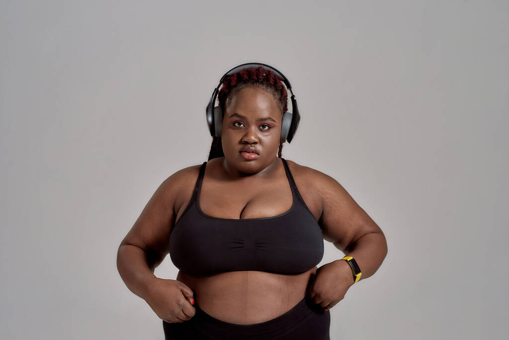Vlastní tvé tělo. Plump, plus velikost africké americké ženy ve sportovním oblečení a sluchátka při pohledu do kamery, stojící ve studiu nad šedým pozadím. Pojetí sportu, zdravý životní styl, tělo pozitivní - Fotografie, Obrázek
