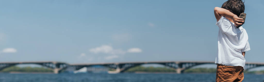 raccolto panoramico di ragazzo che tiene roccia vicino al lago blu - Foto, immagini
