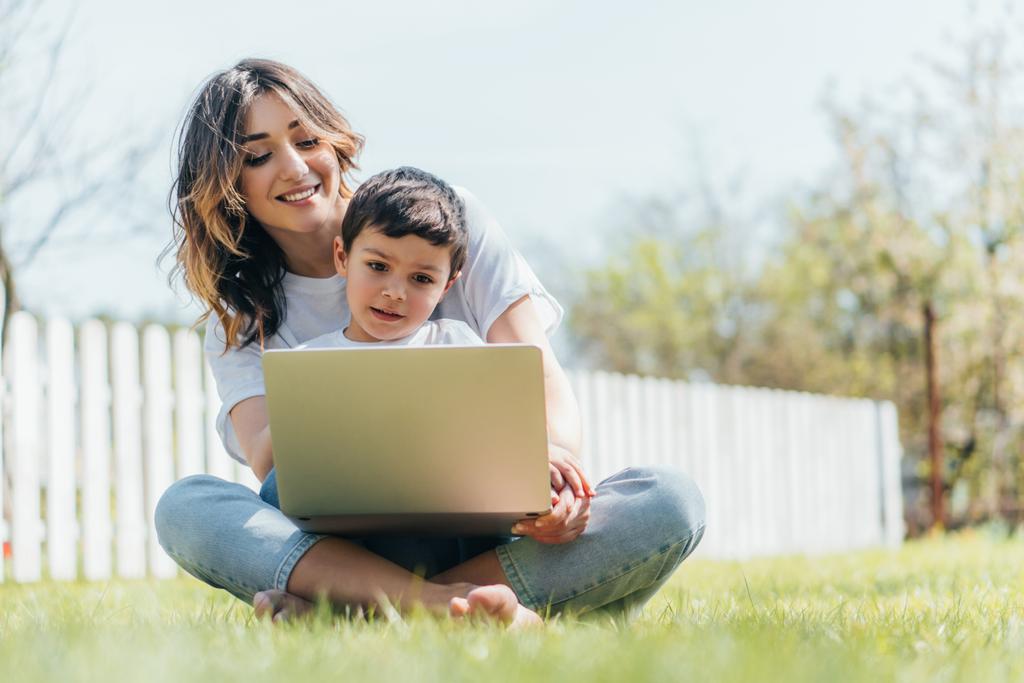 valikoiva painopiste onnellinen äiti ja poika käyttää kannettavaa tietokonetta istuessaan ruohoa  - Valokuva, kuva