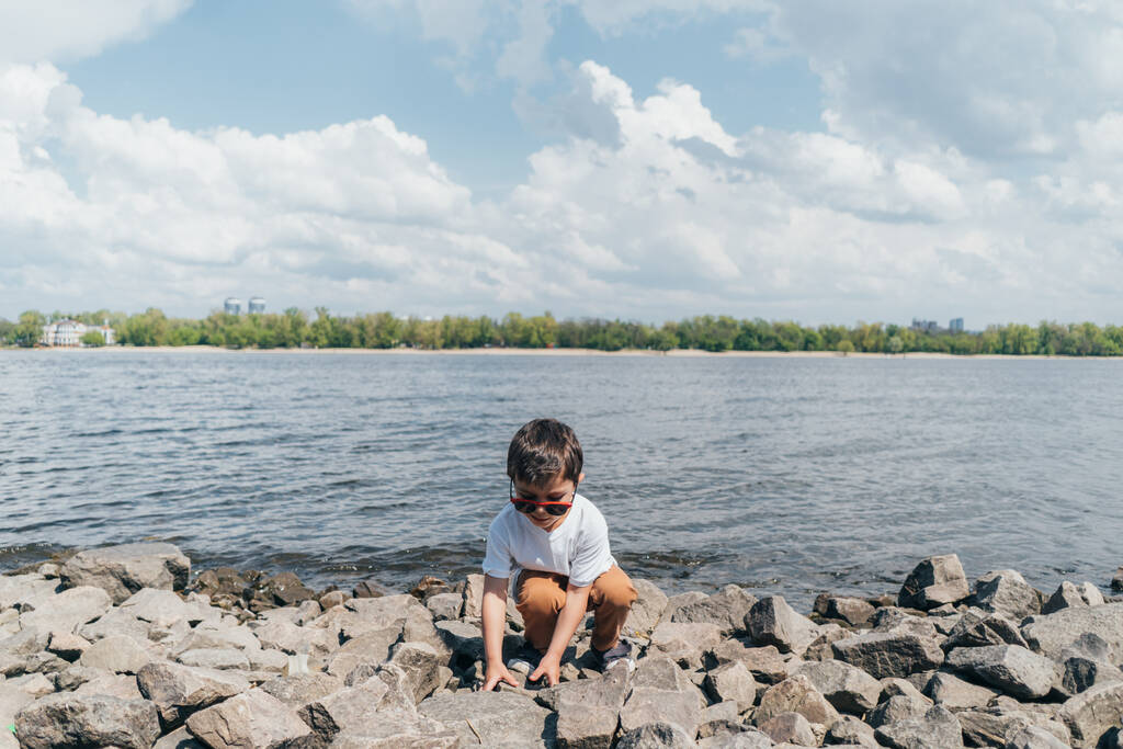 stílusos fiú napszemüvegben megható szikla, miközben ül a tó közelében - Fotó, kép