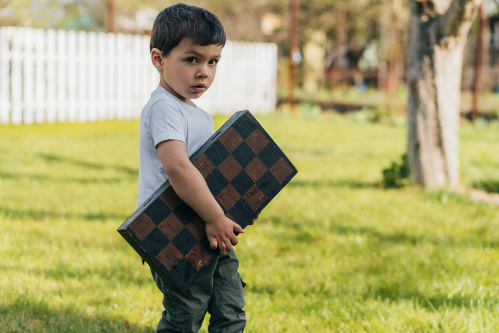 lindo niño sosteniendo tablero de ajedrez y mirando a la cámara
 - Foto, Imagen