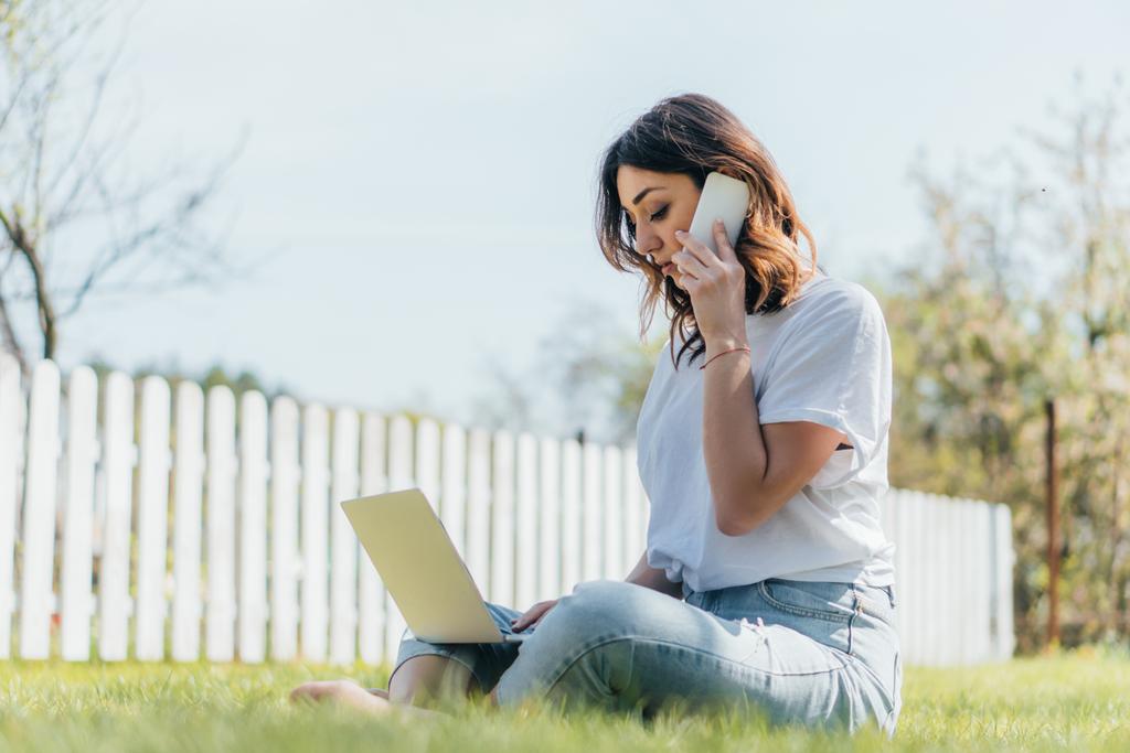 スマートフォンやノートパソコンで草の上に座って話す幸せな女性の選択的な焦点 - 写真・画像